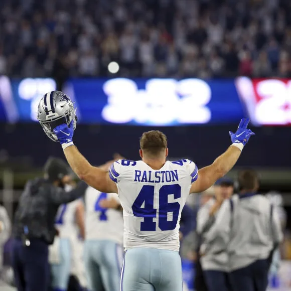 Nick on the sideline during a night game, seen from behind in his number 46 jersey, both arms raised, his helmet held up in one hand, teammates and a full stadium behind him.