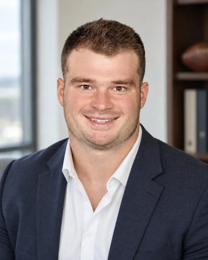 Nick, the founder of AthleteOS, in a navy blazer and an open collar white shirt, smiling, in a daylight office with a football on the shelf behind him.