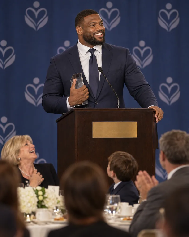 A football player in a navy suit smiles at the lectern of a hotel ballroom as he gives his speech at a children's charity luncheon, a glass award held to his chest and guests at the front table laughing and clapping.