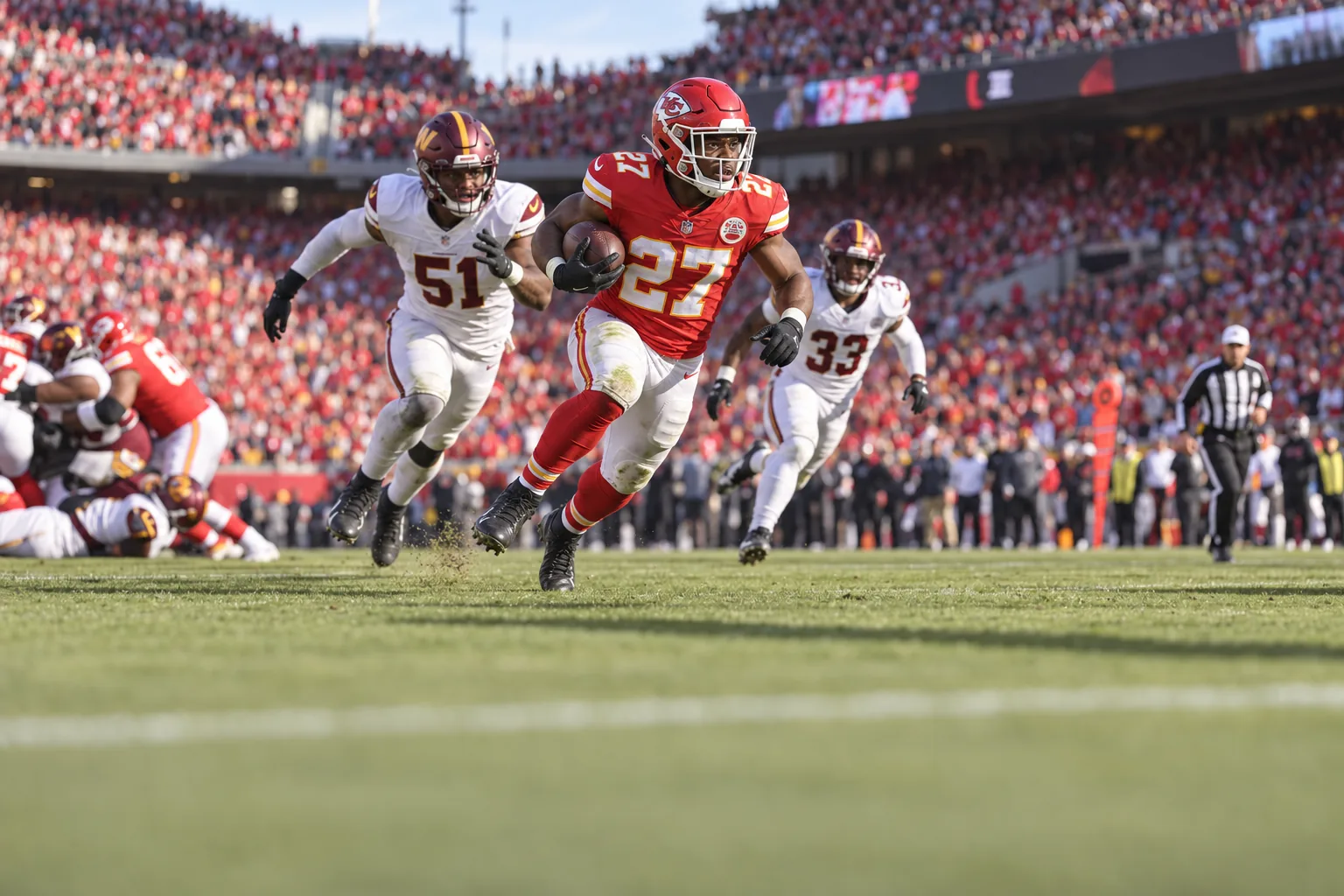 A Kansas City Chiefs running back in a red number 27 uniform cuts upfield with the ball tucked under his arm at a packed Arrowhead Stadium on a clear afternoon, two Washington defenders in white a stride behind him.
