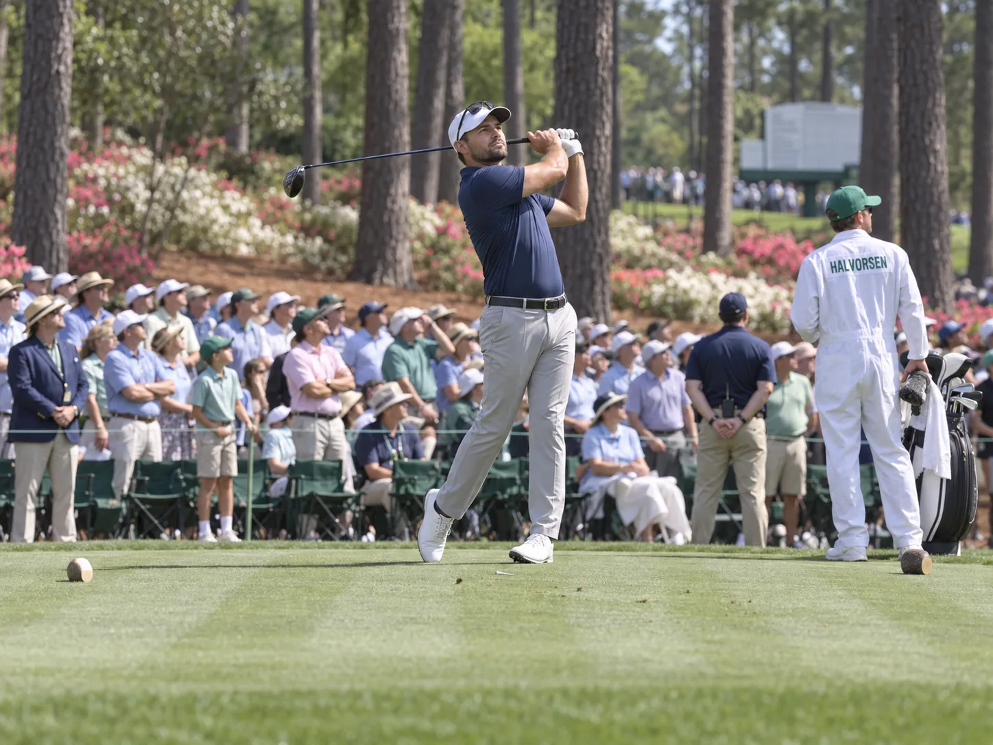 A professional golfer in a navy polo holds his finish on a tee at the Masters, watching his drive, as his caddie in the white tournament coveralls and a gallery under the pines and azaleas follow the ball.